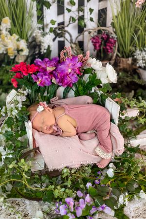 Sleeping baby in pink outfit posed in floral garden setting with colorful blooms.
