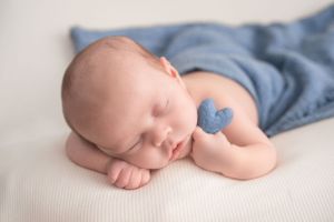 Sleeping newborn in blue wrap with small blue plush toy on white backdrop.