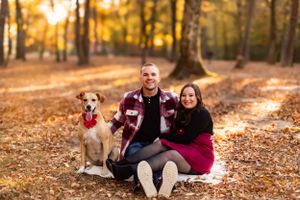 Couple sits with their dog on blanket in autumn forest with golden sunlight.