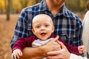 Baby smiles with joy while being held in autumn forest setting.
