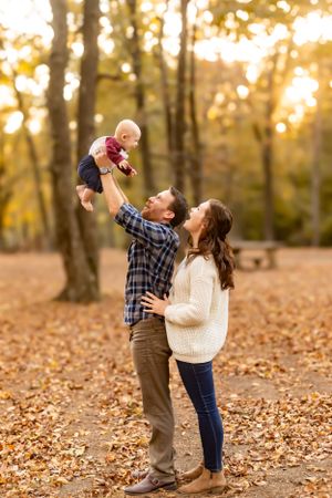 A couple lifts their infant joyfully in a sunlit autumn forest filled with fallen leaves.