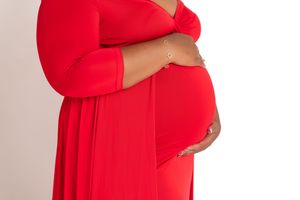 A pregnant woman in a flowing red dress cradles her baby bump against a light background.