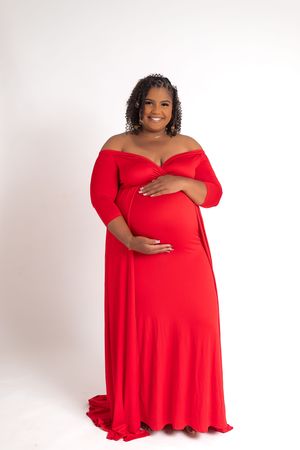 Expectant mother in a flowing red off-shoulder maternity gown poses against a white backdrop with hands on her belly.