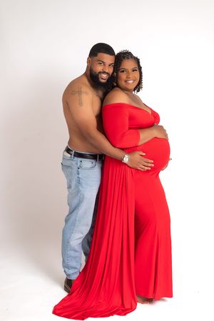 A couple poses together for a maternity photo against a white background, with the woman wearing a flowing red dress.