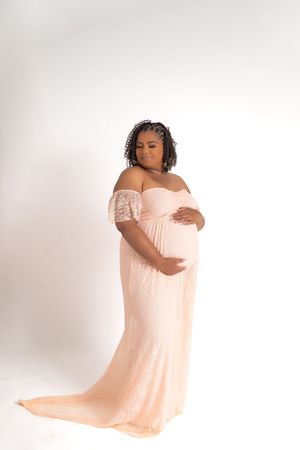 A person in a blush-colored off-shoulder maternity gown poses gracefully against a white backdrop.