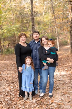 Family poses together on a leaf-covered path in an autumn forest setting.