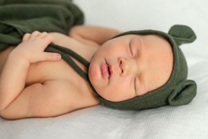 Sleeping baby wearing green bear hat bonnet on white bedding.
