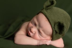 Peaceful newborn sleeping with green bear bonnet against dark green backdrop.