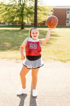 Basketball player in red uniform number 23 and grey skirt poses with ball on sunny outdoor court.