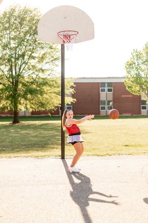 Basketball player in red outfit jumps playfully near outdoor court hoop on sunny day next to brick building.