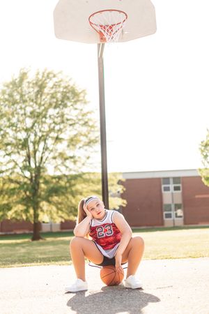 Basketball player in red uniform sits with ball on outdoor court beneath hoop during golden hour.