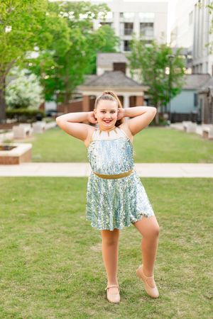 A dancer in a sparkly blue dress performs on a grassy lawn in front of urban buildings on a sunny day.