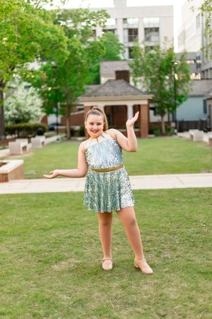 Dancer in sparkly blue dress strikes a pose on grassy lawn with gazebo and city buildings in background.