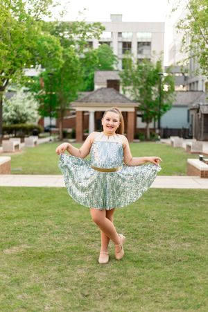 A person in a blue and white floral dress twirls playfully on a grassy lawn in front of an urban building.