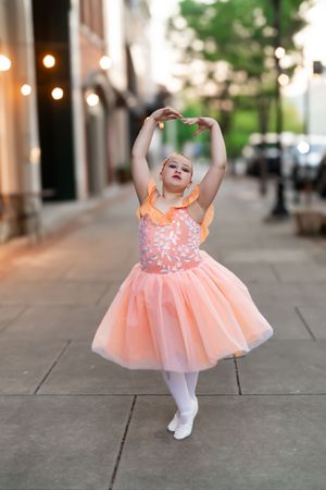 A young dancer in a peachy-pink tutu dress strikes a graceful ballet pose on a city sidewalk with string lights in the background.