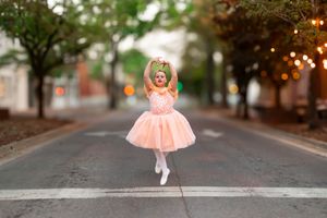 Young ballerina in peach tulle dress performs graceful pose on a tree-lined street with twinkling lights.