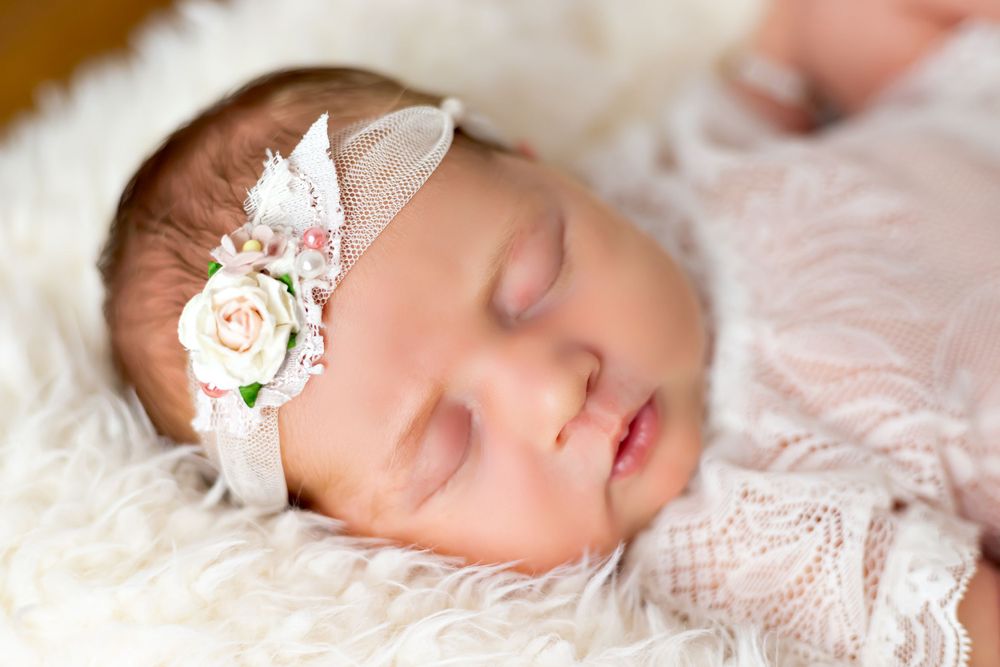 Sleeping baby with delicate floral headband on white lace blanket.