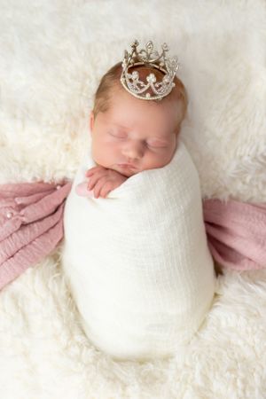 Newborn wrapped in white swaddle wearing silver crown on fluffy cream backdrop.