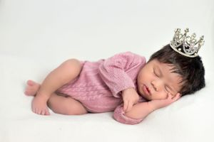 Sleeping newborn in pink outfit wearing silver crown on white background.