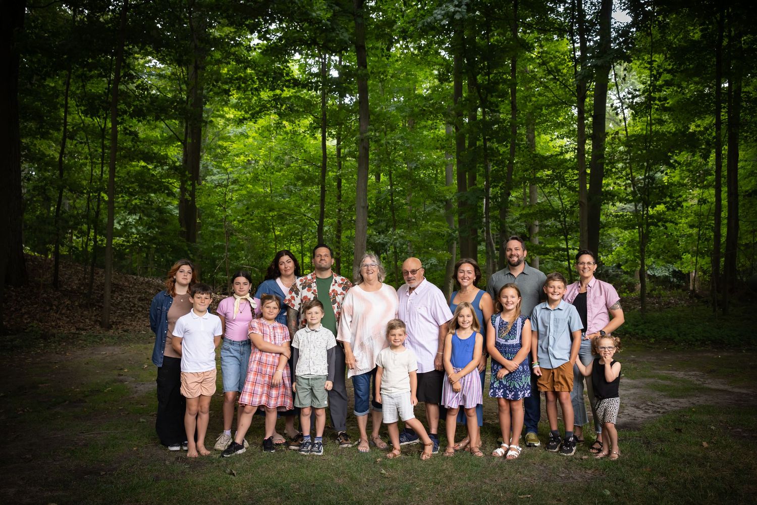 Large family group poses together for a photo in a sunlit forest setting wearing colorful casual clothing.
