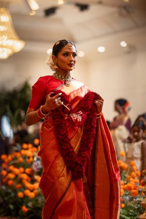 A person in a vibrant red and orange silk saree with traditional Indian jewelry stands among marigold decorations.