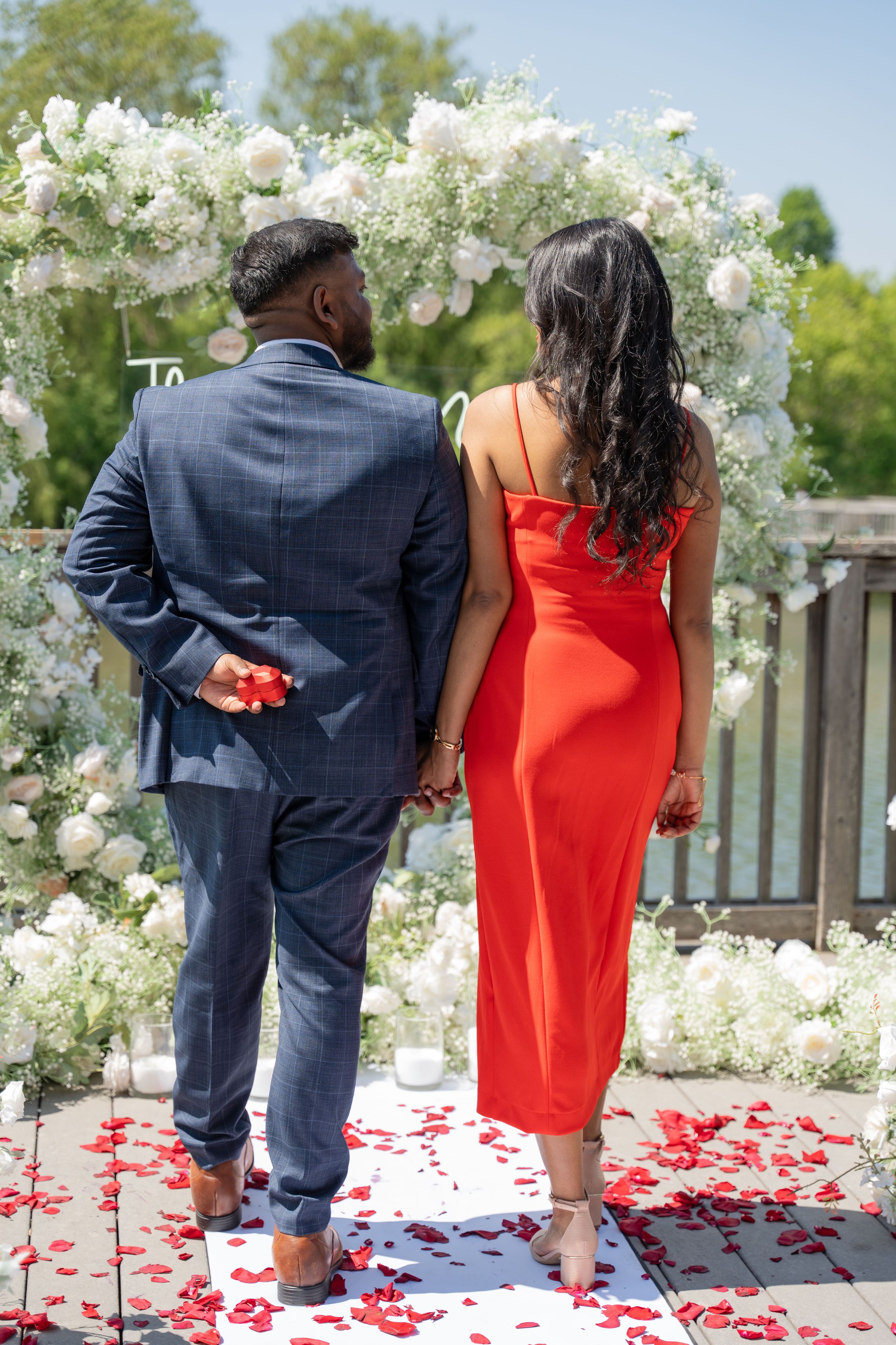 Couple in formal wear poses on white deck with red rose petals and white floral arch.