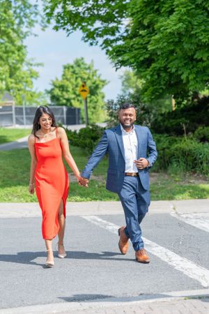 Playful couple in formal attire runs and poses on a paved path surrounded by greenery.