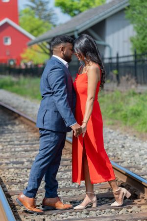 Couple in navy suit and red dress poses playfully on train tracks.
