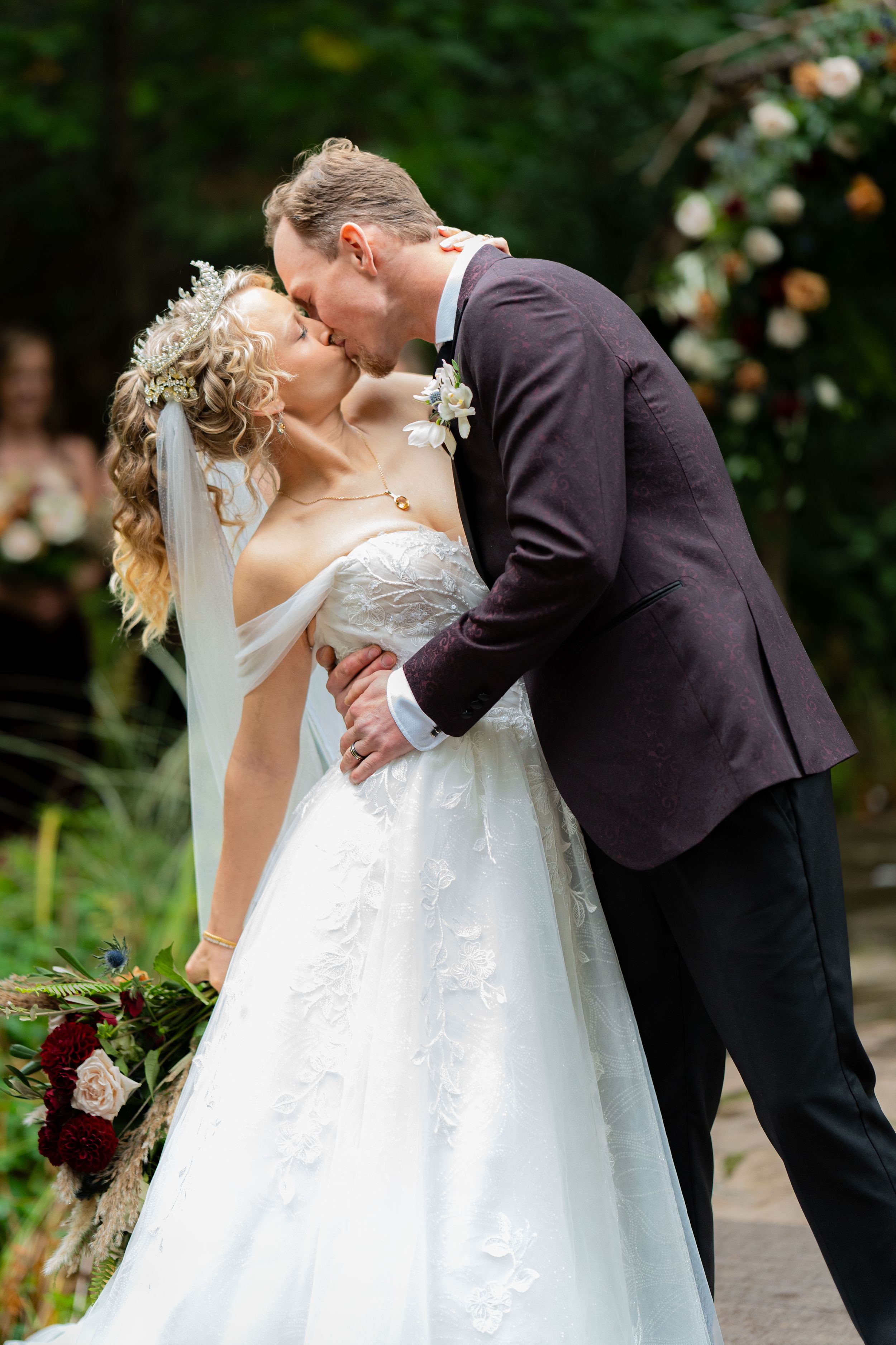 A romantic wedding kiss under a floral arch in a garden setting.