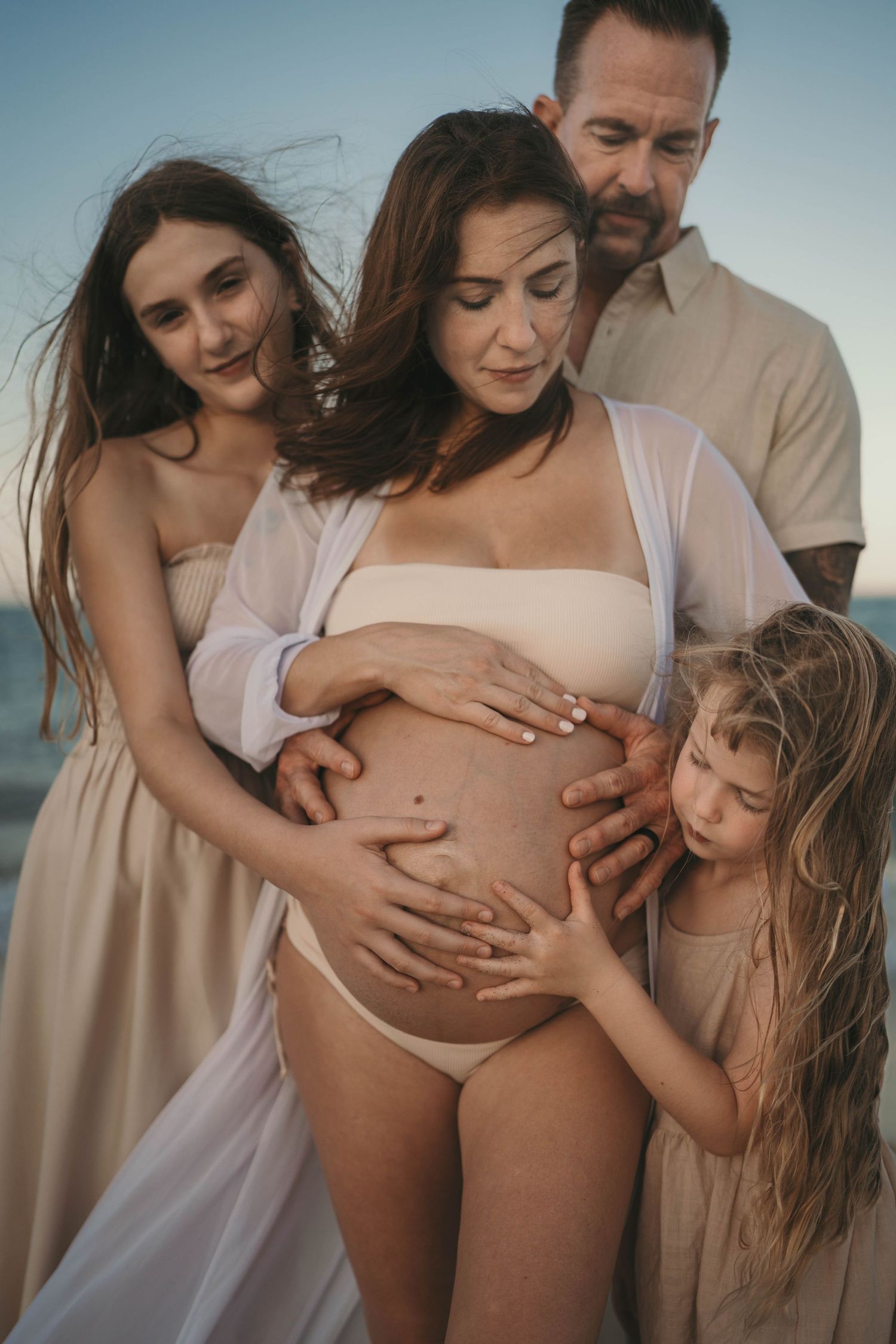Family gathers around to touch pregnant belly on beach at sunset.