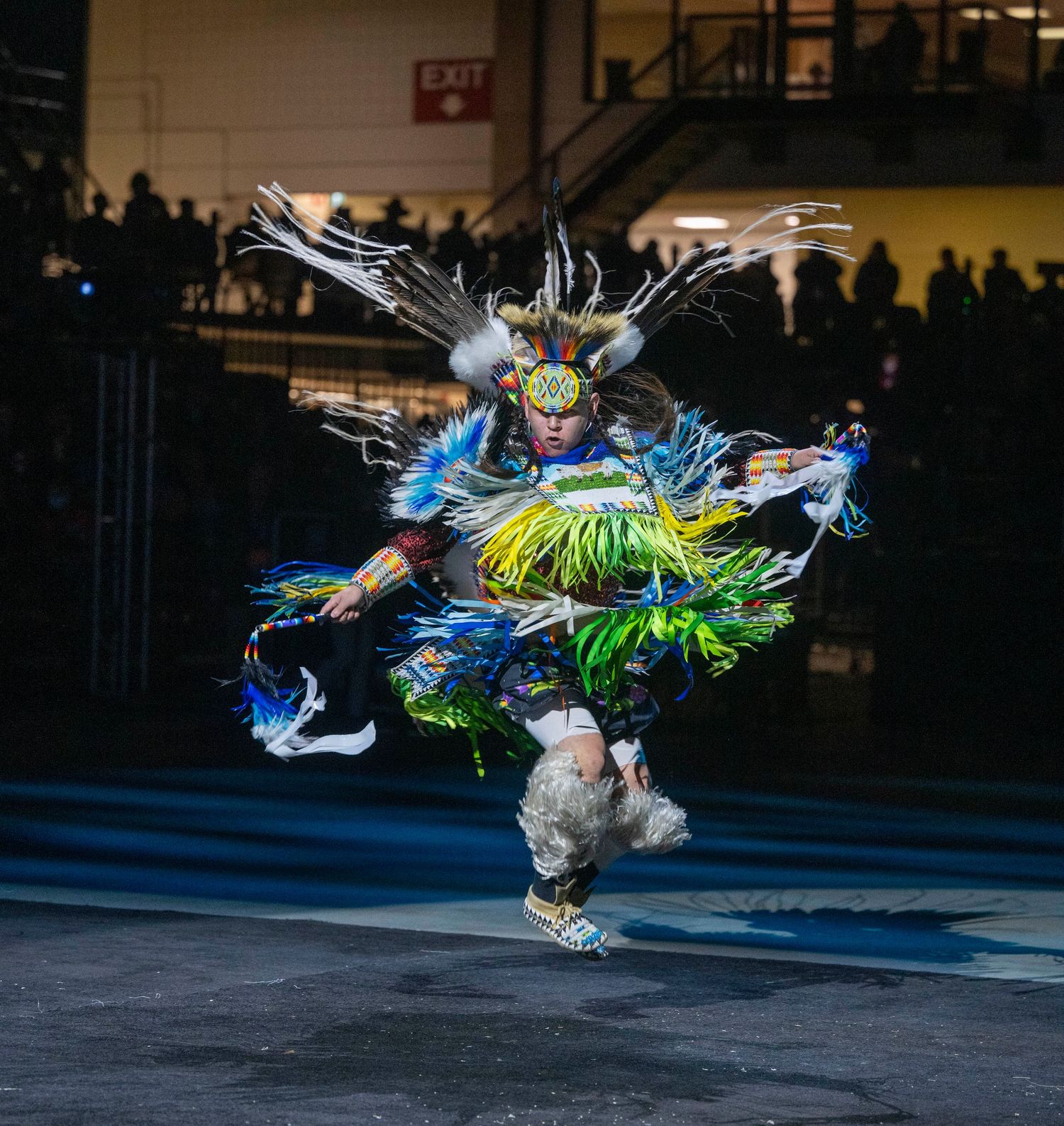 Native American dancer in vibrant blue and green regalia performs a dynamic leap during a traditional powwow performance.