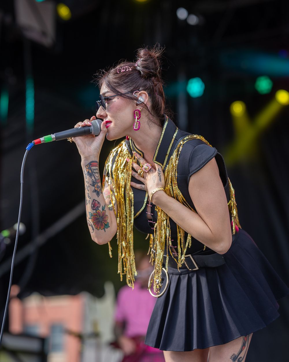 Singer in black dress with gold fringe performs on stage with microphone, showing visible arm tattoos and red earrings.