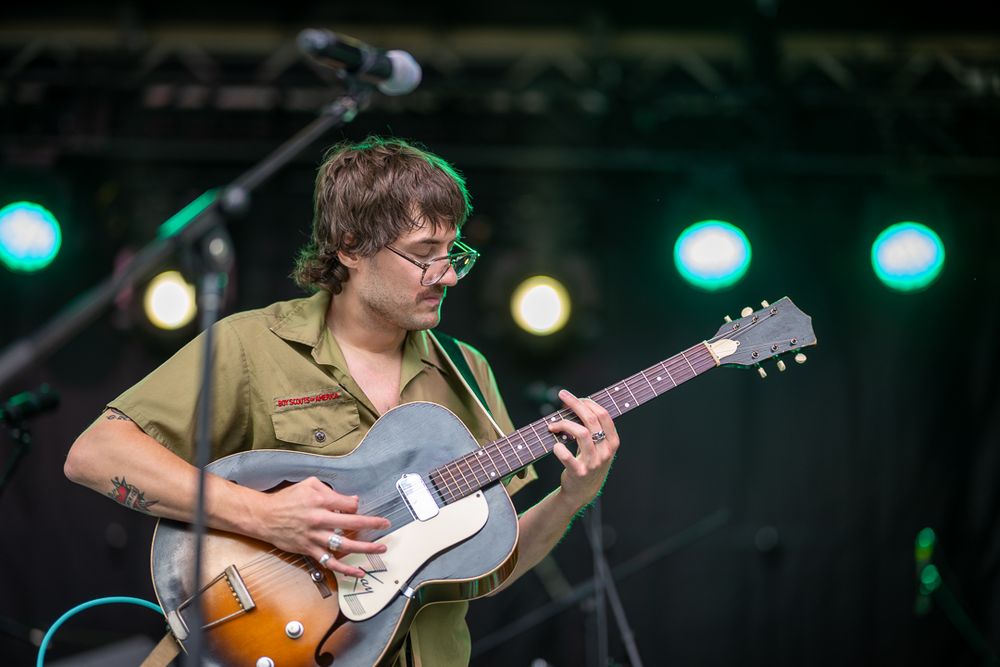 Musician performs on stage with a guitar under green and white stage lighting at a live concert performance.