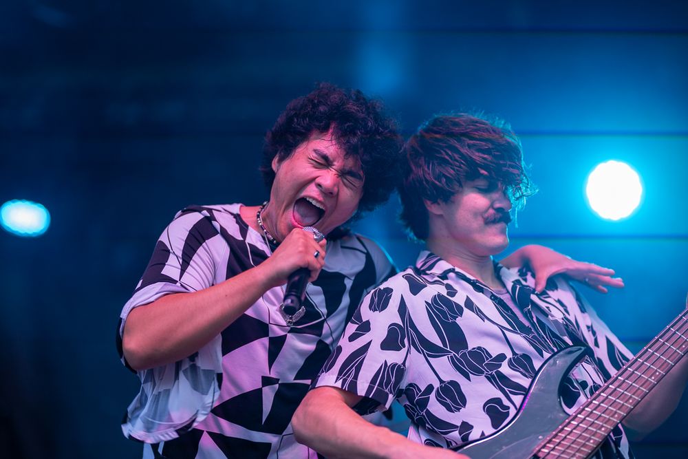 Musicians perform energetically on stage wearing matching black and white patterned shirts under dramatic blue lighting.