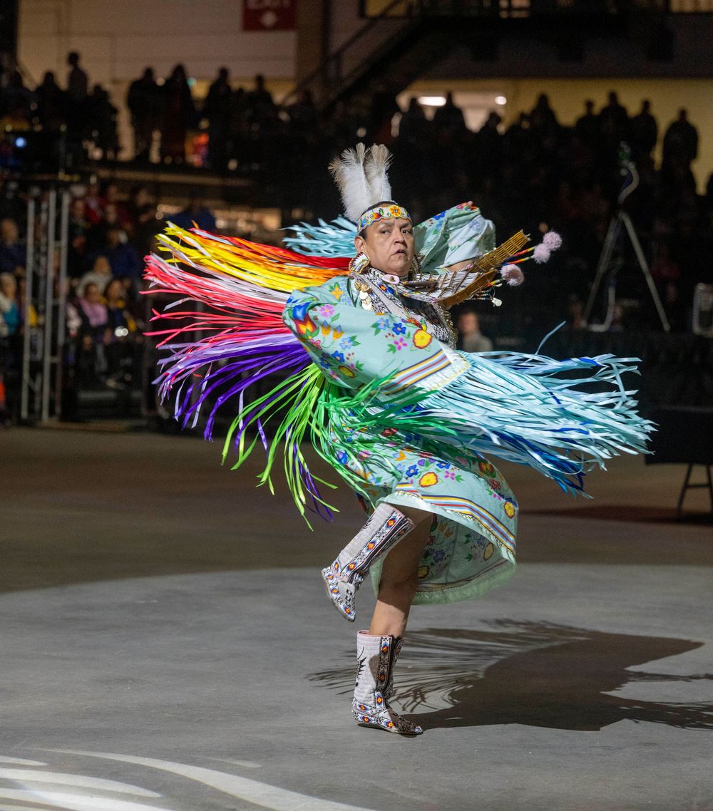 Native American dancer performs traditional dance in vibrant regalia with colorful fringe at indoor powwow event.