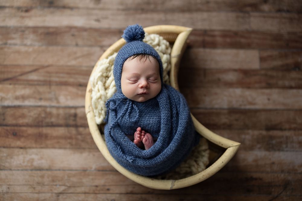 Newborn baby sleeping peacefully in blue knit wrap against rustic wooden backdrop in artistic portrait series.