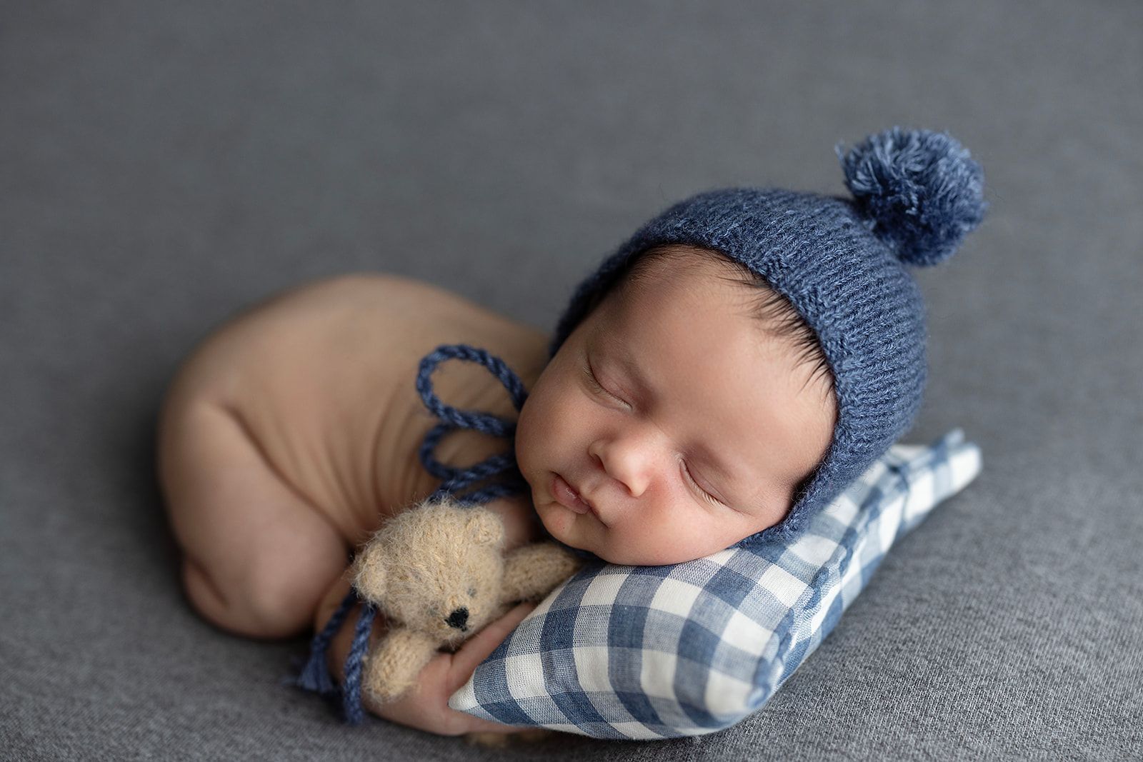 Sleeping newborn in blue and white plaid swaddle lying next to small teddy bear on grey blanket.