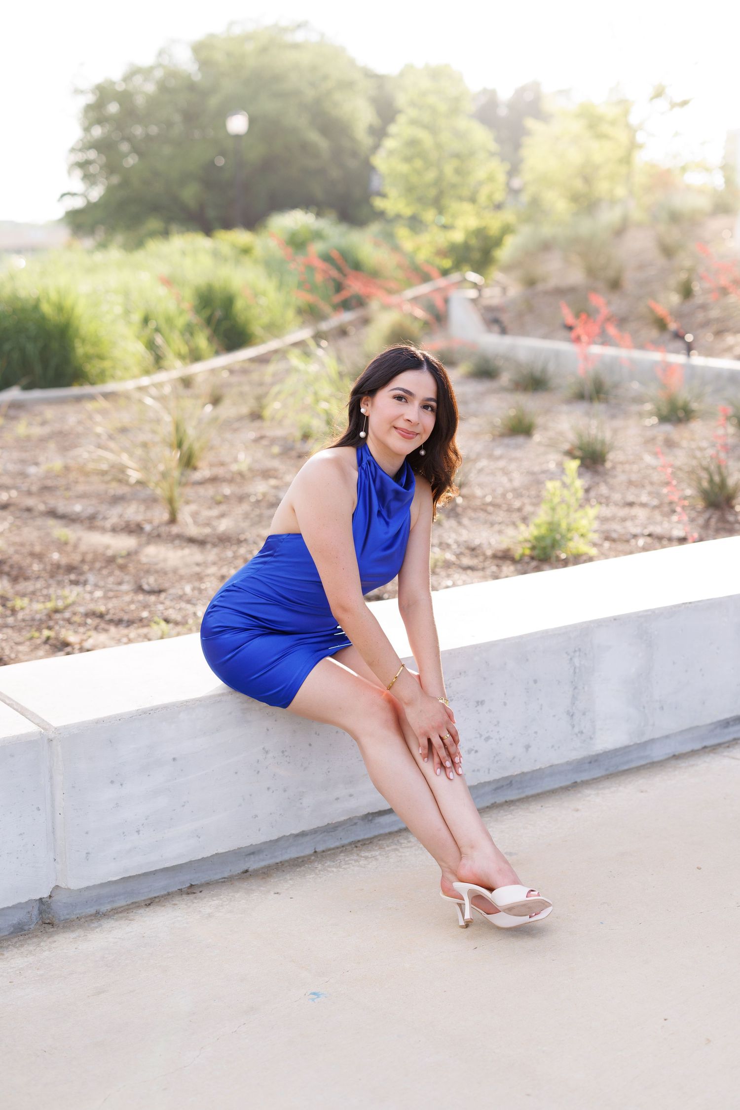 Woman in royal blue dress poses playfully on white concrete ledge outdoors.