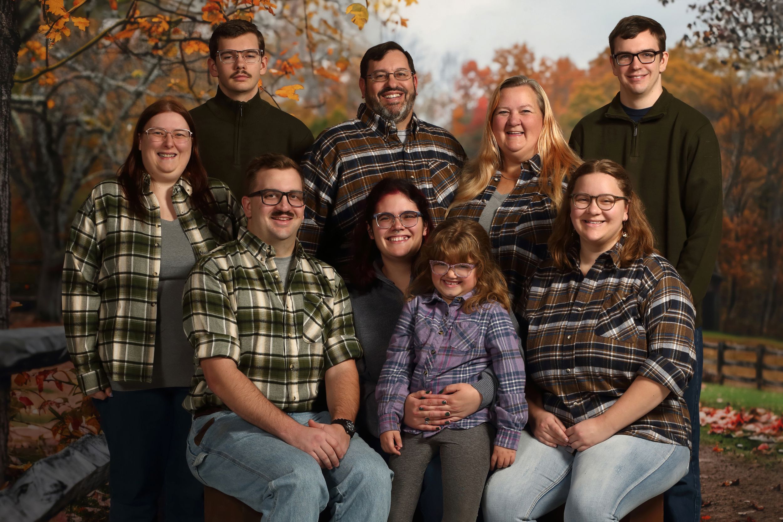 A group of people wearing plaid and casual clothing pose together outdoors during autumn with fall foliage in background.