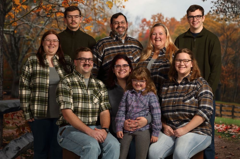 A group of people wearing plaid and casual clothing pose together outdoors during autumn with fall foliage in background.