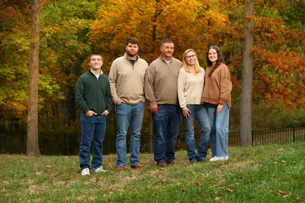 A group of five people stand together on a grassy hill with vibrant autumn foliage in the background.