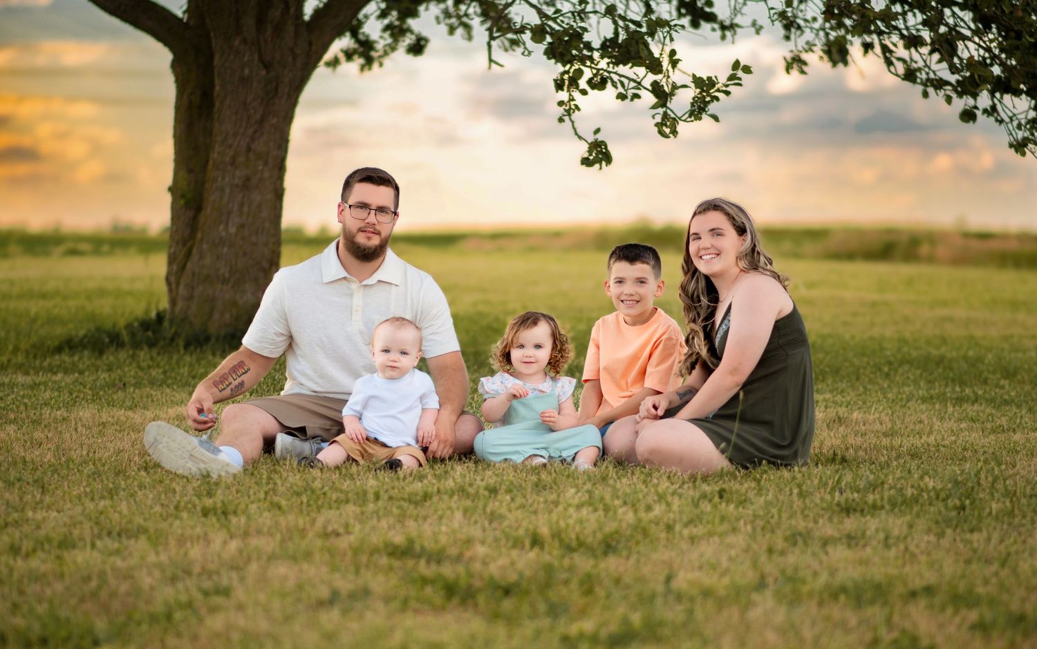 Happy family of five sitting together under a large tree at sunset on green grass.