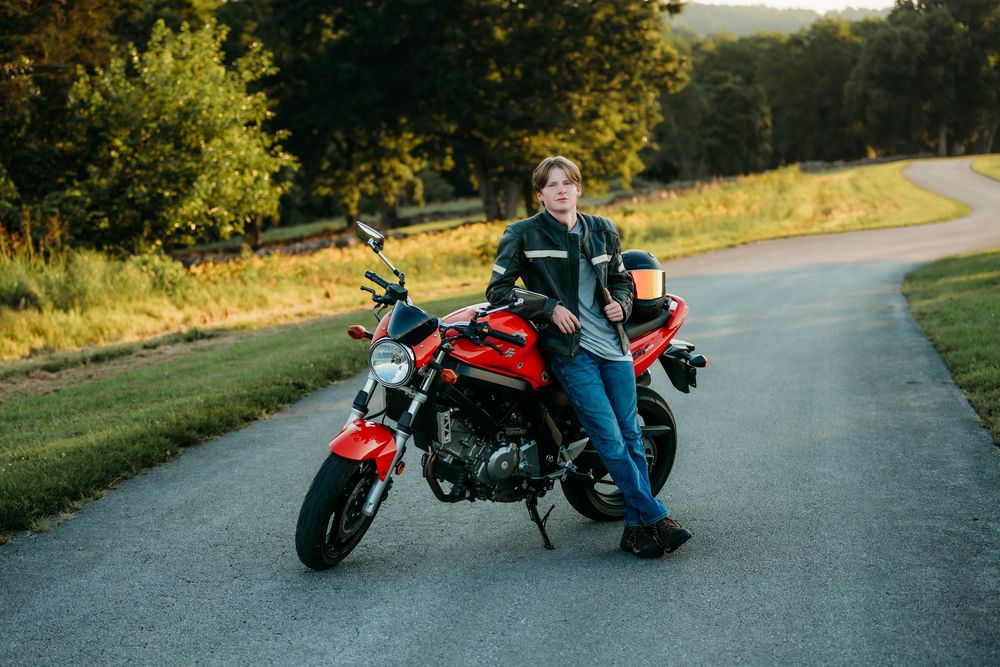 Young person in jeans and jacket leaning against a red Ducati motorcycle on a quiet road surrounded by green trees.