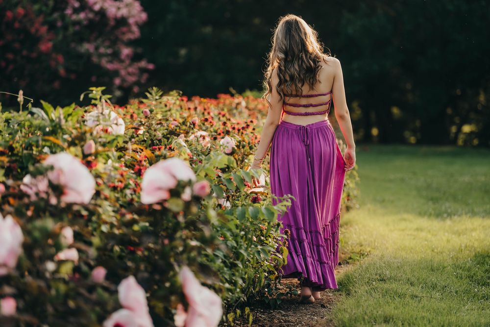 Woman in purple maxi skirt and crop top walking through a lush garden with colorful blooming flowers.
