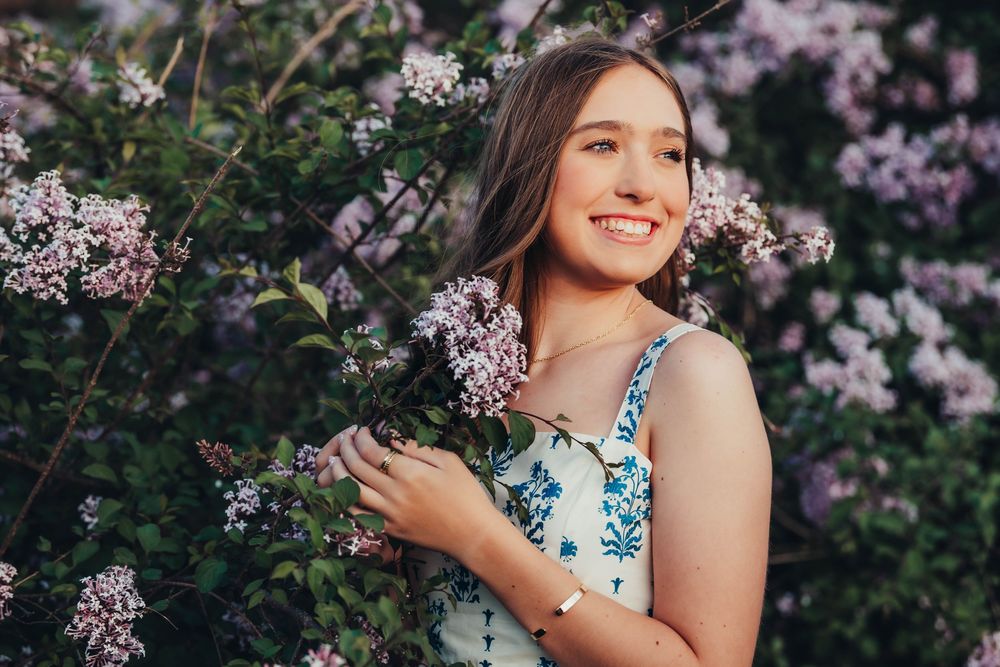 Smiling young woman in floral dress holding lilac blooms outdoors in a garden setting.