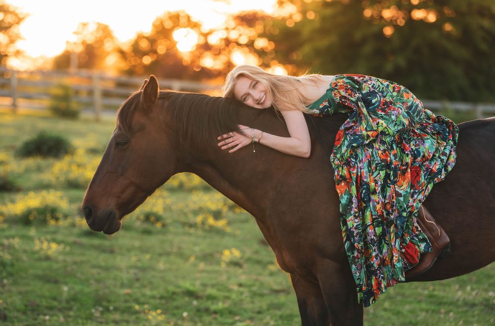 Blonde woman in floral dress leaning on brown horse at golden sunset in green pasture.