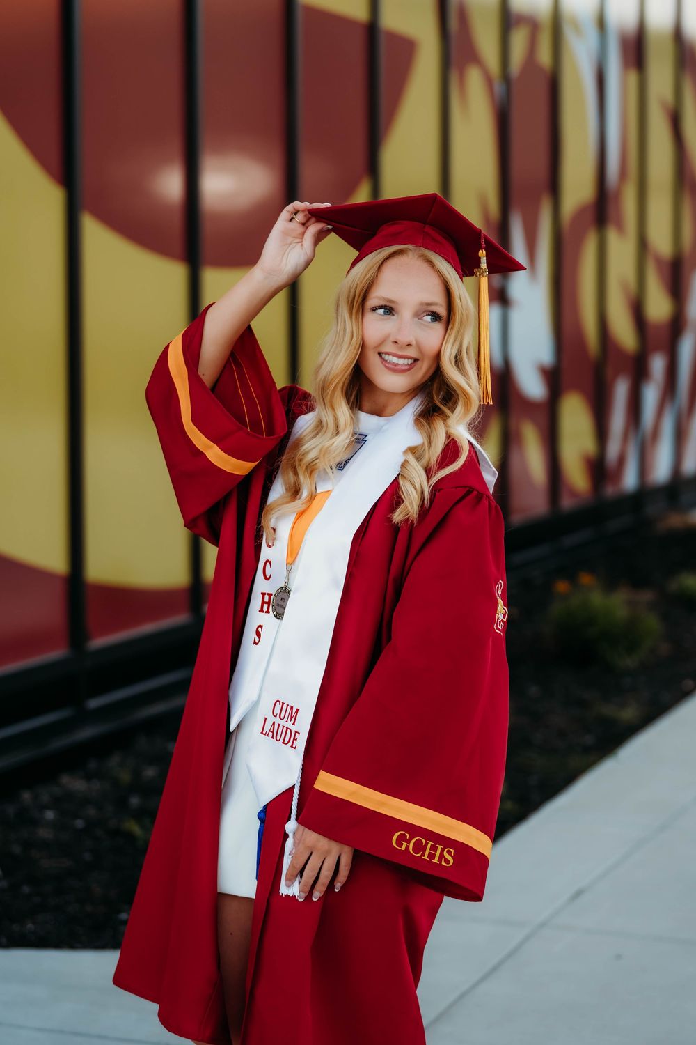 Smiling female graduate in red cap and gown with cum laude sash poses outdoors at GCHS graduation ceremony.