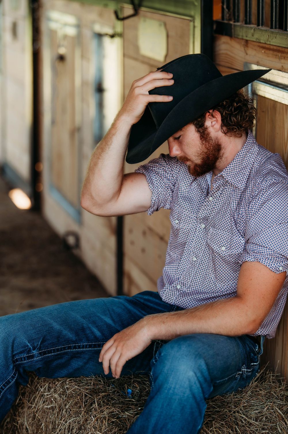 A person in western attire adjusts their black cowboy hat while sitting on hay bales in a rustic barn setting.