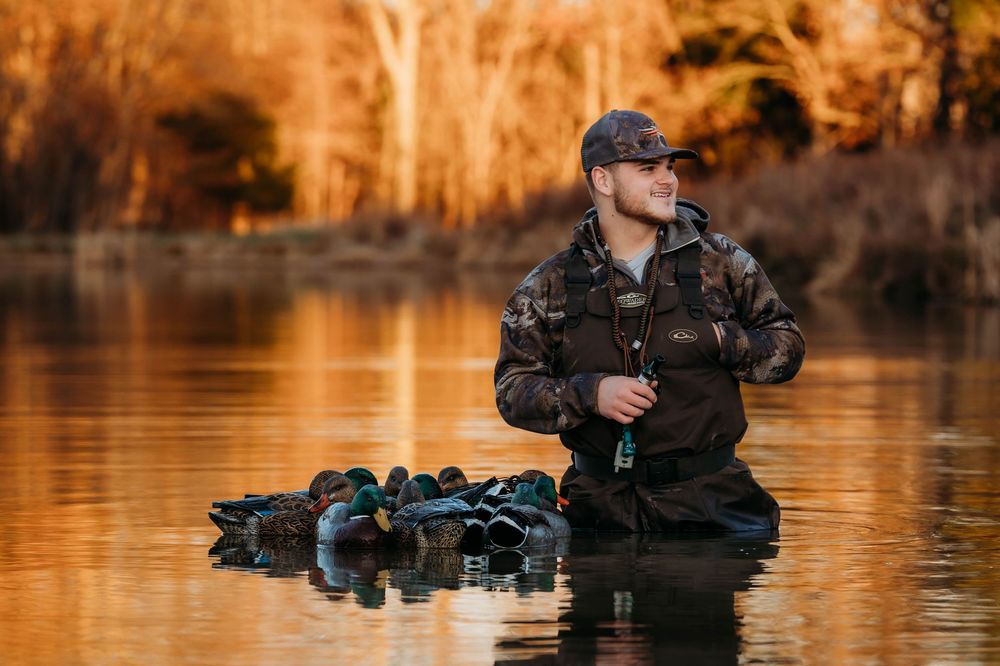 Professional photographer wades through tranquil water at sunset while carrying camera equipment.