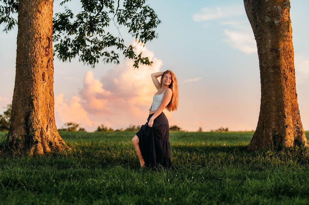 Someone in a maxi dress poses between two large trees at sunset in a grassy field during golden hour.